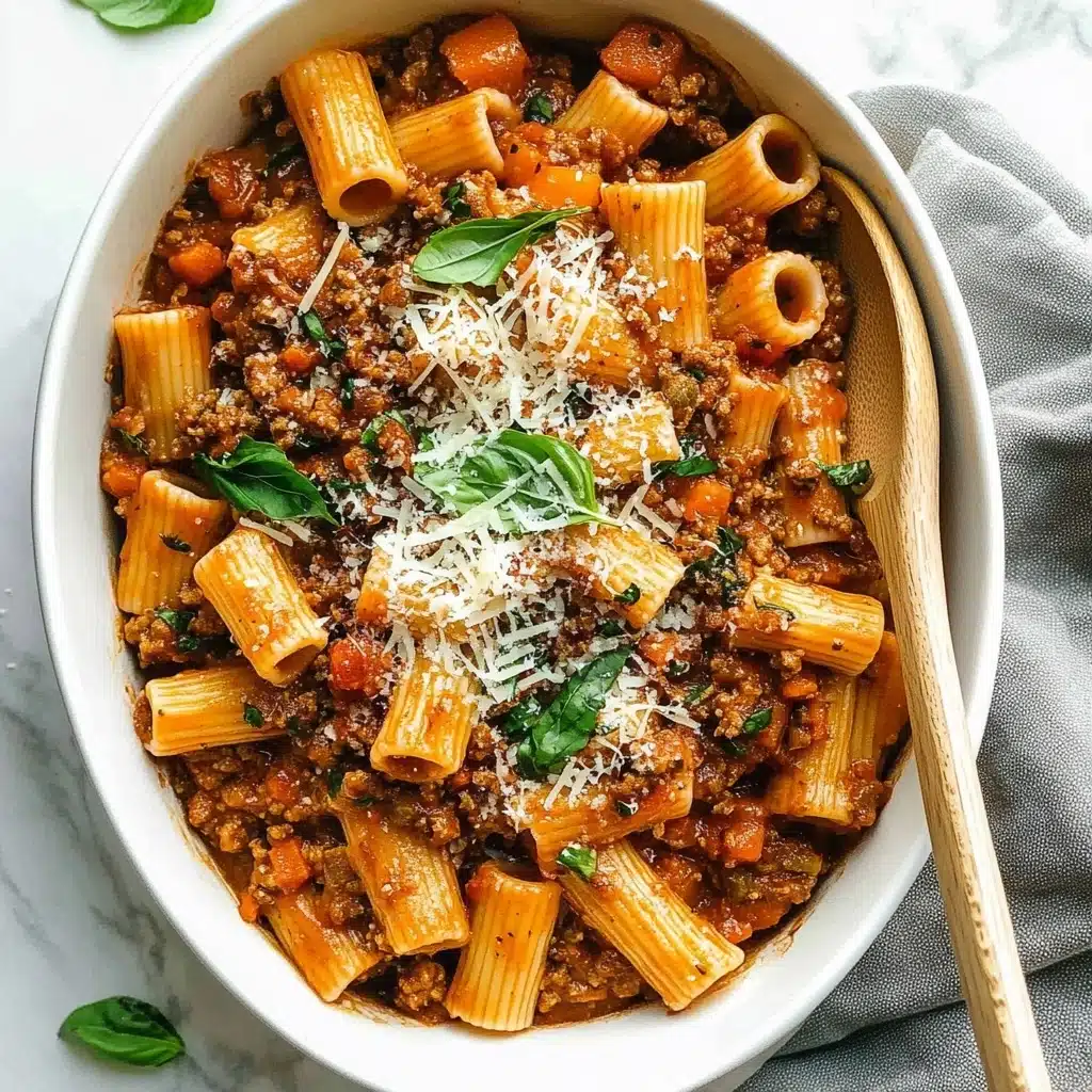 Spicy Sausage Rigatoni, spicy sausage pasta recipe, easy spicy rigatoni, hearty sausage dinner, flavorful pasta dish A white oval dish filled with rigatoni pasta mixed evenly with a thick brown meat sauce containing small pieces of carrot and tomato. The orange pasta tubes have ridges and are scattered around the dish with the sauce layered throughout. On top, finely shredded white cheese is sprinkled, adding a delicate texture, along with thin strips of fresh green basil leaves spread over the dish. A wooden spoon with a white handle is placed partially inside the dish on the right side. The dish sits on a white marbled surface with a textured gray cloth to the right.