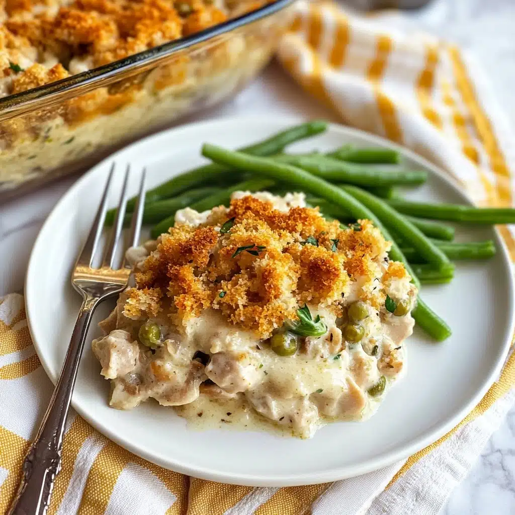 Chicken Stuffing Bake, Easy Chicken Casserole, Comfort Food Dinner, Hearty Chicken and Stuffing, Family-Friendly Chicken Bake A white plate holds a serving of a creamy dish with three visible layers: a melting white sauce base, a thick middle layer of tender light beige poultry, and a top layer of golden-brown, chunky bread crumb topping with herbs. Next to the main dish, there is a side of bright green steamed green beans. The plate is placed on a white and mustard yellow checkered napkin over a white marbled texture surface. In the background, there is a clear baking dish with more of the creamy breadcrumb-topped dish visible. A silver fork lies near the top edge of the plate.