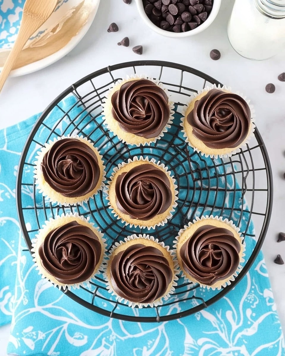 Eight vanilla cupcakes with smooth, dark brown chocolate frosting piped in a swirl resembling a rose, placed on a black round wire cooling rack. Each cupcake shows two layers: the light golden cake base and the glossy chocolate frosting on top. The rack sits on a bright blue fabric with a white floral pattern, which is on top of a white marbled surface. Nearby, there is a white plate with an intricate design and two wooden utensils on the top left, a small white bowl filled with dark chocolate chips at the top right, and a clear bottle with milk and a white straw with blue dots on the far right. Small chocolate chips are scattered around on the marbled surface. The photo taken with an iphone --ar 4:5 --v 6.1