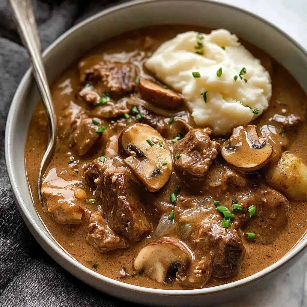 Slow Cooker Beef Stroganoff, Beef Stroganoff, Crockpot Beef Stroganoff, Comfort Food Recipes, Easy Slow Cooker Dinners A deep bowl filled with thick, creamy brown sauce, containing several pieces of browned meat and sliced mushrooms, all covered in the sauce with small green chives sprinkled on top. There is a dollop of white mashed potatoes partially covered by sauce near the back of the bowl. A silver spoon rests on the left side of the bowl, which is placed on a soft dark gray fabric against a white marbled texture.