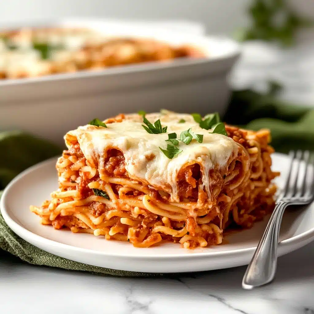 Baked Spaghetti with Cream Cheese, cheesy baked spaghetti, creamy pasta bake, simple baked spaghetti recipe, comfort food spaghetti A square slice of baked pasta layered with cooked spaghetti in reddish tomato sauce mixed with small pieces of green herbs, topped with melted white cheese and small bits of tomato, served on a white plate with a silver fork beside it, placed on a green cloth on a white marbled surface; in the background, a white dish with more pasta is blurred.