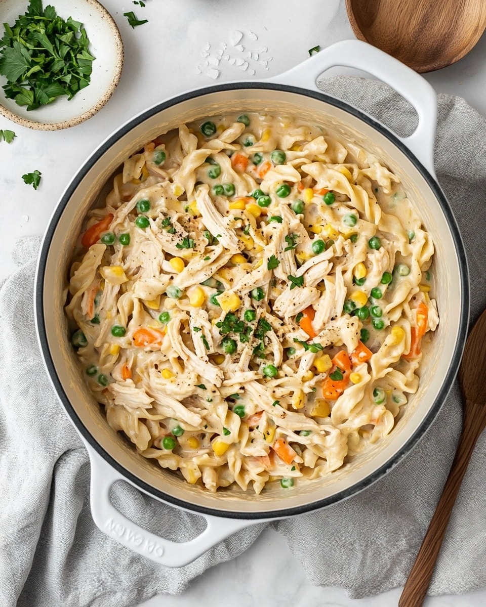 Chicken Pot Pie Pasta, easy creamy chicken pasta, quick comfort food, hearty pasta dishes, homemade dinner ideas A white pot filled with creamy pasta that has three main layers: pale yellow twisted noodles, cooked shredded chicken mixed throughout, and colorful vegetables including orange carrot slices, green peas, and yellow corn kernels. The creamy sauce covers all the ingredients and is sprinkled with green herbs on top. The pot is placed on a crumpled gray cloth over a white marbled surface. Nearby, there is a white plate with a simple textured design, a small wooden bowl filled with white salt and a wooden spoon, a small dish with green herbs, and two silver forks.