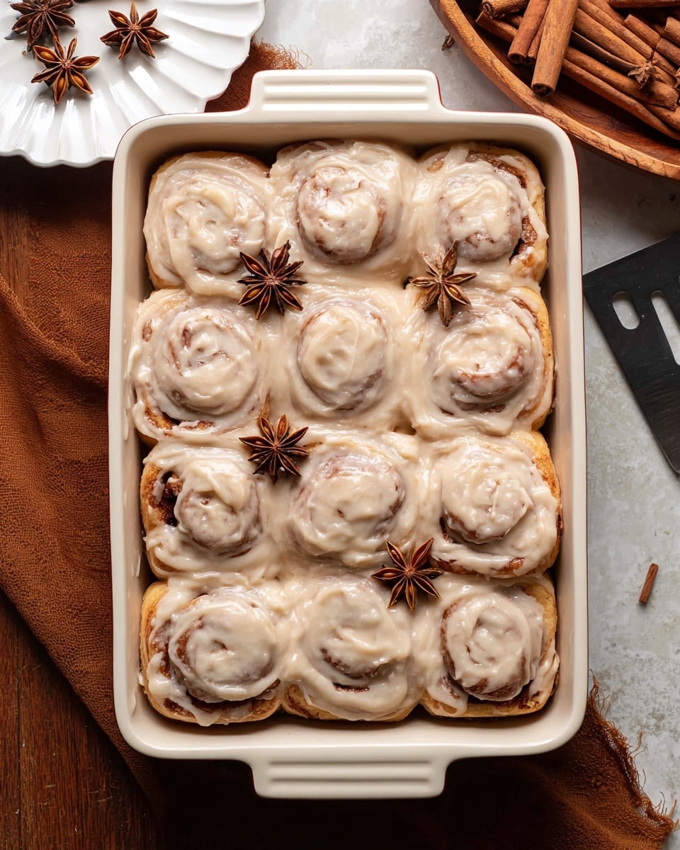 A white rectangular dish contains twelve cinnamon rolls arranged in three rows of four. Each cinnamon roll is thickly covered with creamy light brown icing that has a smooth, swirled texture following the spiral shape of the rolls. The cinnamon rolls show hints of golden brown dough beneath the icing. Three star anise pieces are placed on top for decoration. The white marbled surface beneath the dish contrasts with the wooden bowl of cinnamon sticks and star anise in the background. Nearby, a white scalloped plate and a dark metal serving spatula are visible. photo taken with an iphone --ar 4:5 --v 7