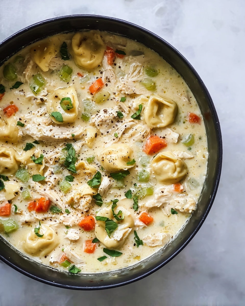 A black pot filled with creamy chicken noodle soup is shown from above on a white marbled texture. The soup has three main layers: a pale yellow creamy broth base with thick texture, pieces of shredded white chicken mixed throughout, and small diced orange carrots and light green celery scattered evenly. There are small tortellini pasta pieces, round with a slight hollow center, floating in the soup. Fresh green herb leaves sprinkled on top add a bright color contrast, and a light dusting of ground black pepper is visible across the surface. photo taken with an iphone --ar 4:5 --v 7