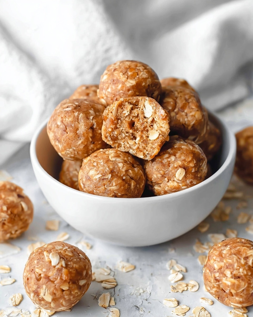 A white bowl filled with round, oatmeal energy bites that are light brown with visible oat flakes throughout. One energy bite is placed on top, broken in half to show a soft, crumbly texture with small pieces of oats and nuts inside. Several more energy bites are scattered around the bowl on a white marbled surface, and a white cloth is draped in the background. The overall look is warm and inviting, focusing on the natural texture and color of the bites. photo taken with an iphone --ar 4:5 --v 7