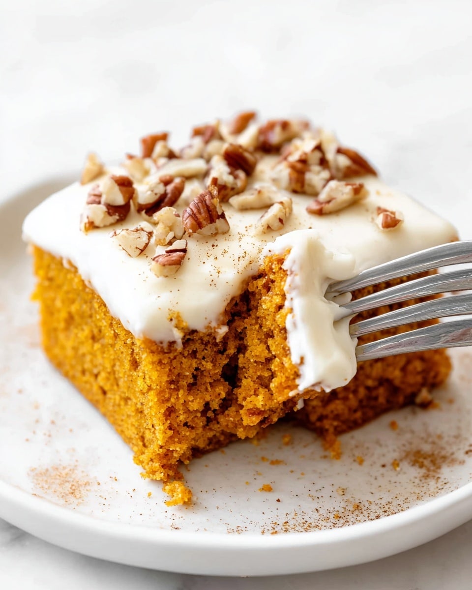 A square piece of orange-brown moist cake with a soft, airy texture sits centered on a white plate. On top of the cake is a thick layer of smooth white frosting, sprinkled lightly with brown spice powder, and decorated with chunks of chopped pecans scattered across the surface. The plate rests on a white marbled surface, with a silver fork placed near the bottom left corner. In the blurred background, another white plate with more cake and a small orange pumpkin add to the autumn theme. Photo taken with an iphone --ar 4:5 --v 7