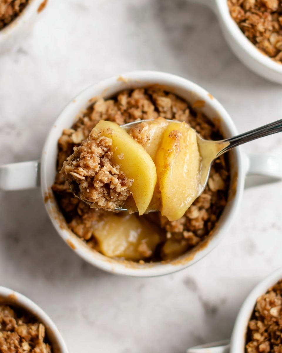 Three white mugs are filled with apple crisp, each showing two main layers: at the bottom, soft slices of light yellow and green apples, partially hidden by a top layer of golden brown oat crumble with a rough texture. One mug has two scoops of pale beige ice cream melting slightly into the crisp. The mugs are placed on a white marbled surface with scattered apple slices and a pale green cloth with spoons beside them. Photo taken with an iphone --ar 4:5 --v 7