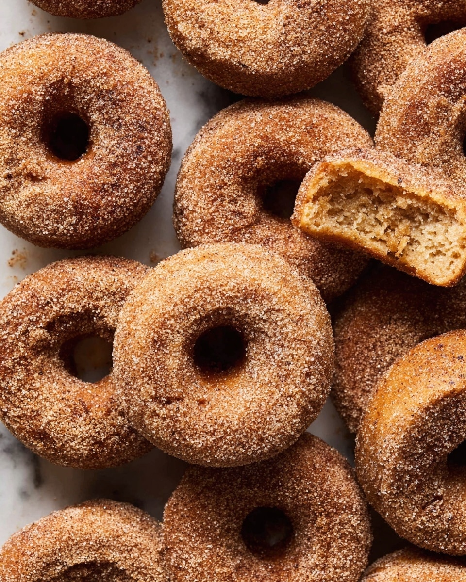 A close-up top view of a group of small round donuts with a hole in the center, all covered evenly in a light brown sugar coating giving a rough texture. The donuts are touching each other, filling the entire frame, with one donut at the top right having a piece broken off showing the soft, airy inside in a light golden brown color. The donuts have a warm, slightly darker brown baked surface visible under the sugar layer. They rest on a white marbled texture background. photo taken with an iphone --ar 4:5 --v 7
