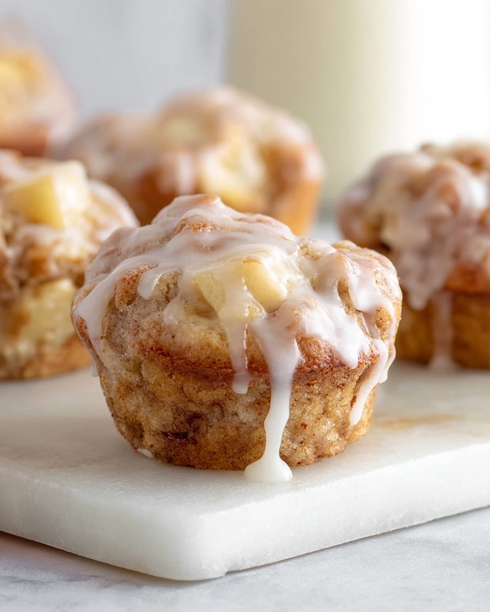 A close-up image shows a woman's hand holding a small, bitten piece of a light brown muffin with a moist texture, revealing a soft, pale green apple chunk inside. The muffin has a thin white icing layer on top, slightly dripping on the edges. In the blurred background, a white plate holds more muffins, and there are faint shapes suggesting a glass bottle and a glass filled with milk on a white marbled surface. The lighting is soft and natural, highlighting the muffin’s crumb texture. photo taken with an iphone --ar 4:5 --v 7