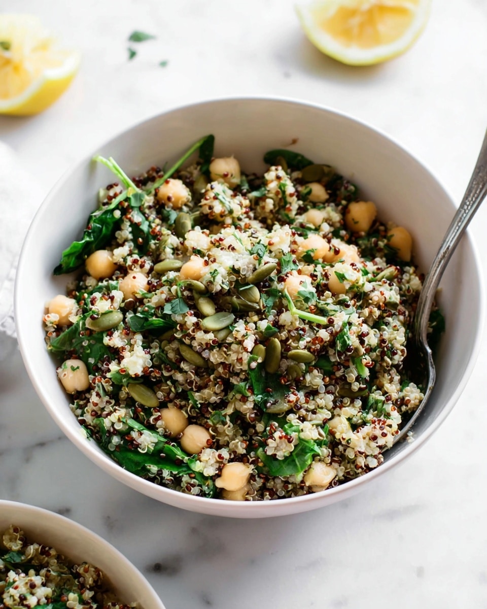 A white bowl filled with a quinoa salad showing about three main layers: the base layer is white and red quinoa with small round seeds, mixed with pale beige chickpeas; the middle layer has bright green leafy herbs and spinach pieces scattered throughout; the top layer shows small green pumpkin seeds and finely chopped herbs sprinkled on top. The bowl is set on a white marbled surface with a silver spoon resting inside the bowl and two lemon halves slightly out of focus in the background. Photo taken with an iphone --ar 4:5 --v 7