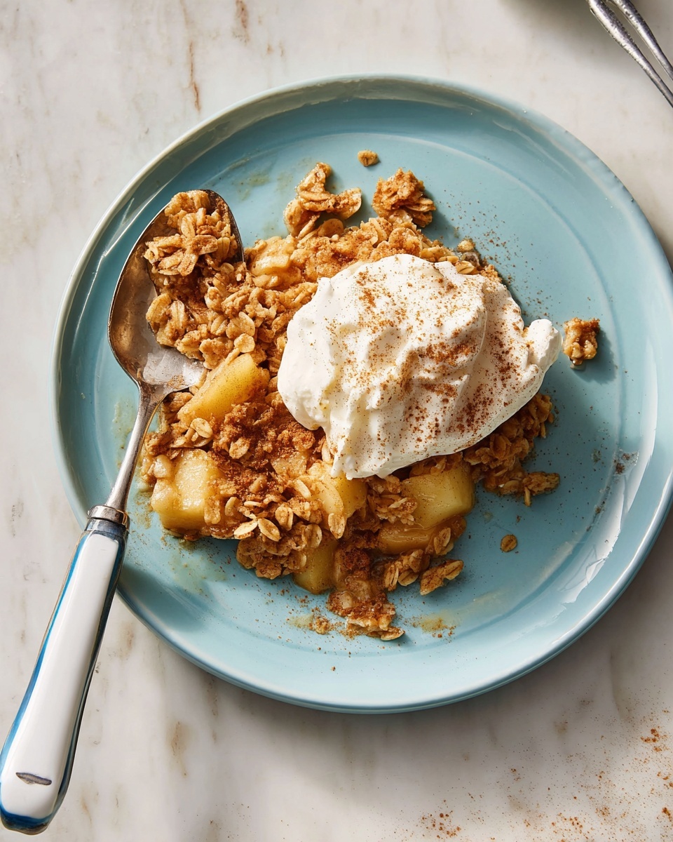 On a white plate, there is a serving of oatmeal apple crumble with visible chunks of soft apple and a golden-brown oat topping that looks crunchy and crumbly. On top of the crumble, there is a dollop of white cream sprinkled with light brown cinnamon powder. A silver spoon rests on the upper part of the plate, slightly touching the crumble. The surface beneath the plate is a white marbled texture. photo taken with an iphone --ar 4:5 --v 7