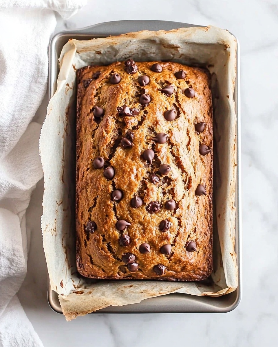 A loaf of golden brown baked bread with chocolate chips scattered all over the top, sitting inside a silver metal baking pan lined with light brown parchment paper on two sides; the bread surface is slightly cracked, showing a moist and soft texture beneath the crunchy top. The pan rests on a white marbled surface, with part of a white cloth towel visible on the left. Photo taken with an iphone --ar 4:5 --v 6.1