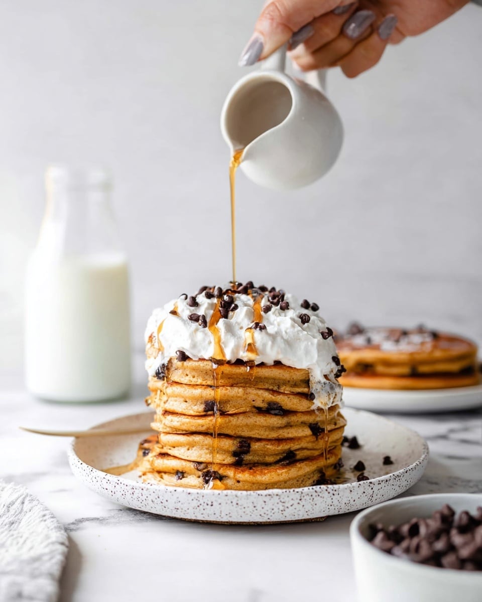 A stack of seven thick, round pumpkin pancakes with visible small chocolate chips layered evenly in each pancake sits on a white speckled plate. The pancakes are golden orange with slightly browned edges, and amber syrup drips down the sides in thin streams. On the top pancake, there is a dollop of white whipped cream sprinkled with mini chocolate chips and a light dusting of cinnamon. The scene is set on a white marbled surface with blurred silverware in a glass jar and a bottle of milk in the background. photo taken with an iphone --ar 4:5 --v 7