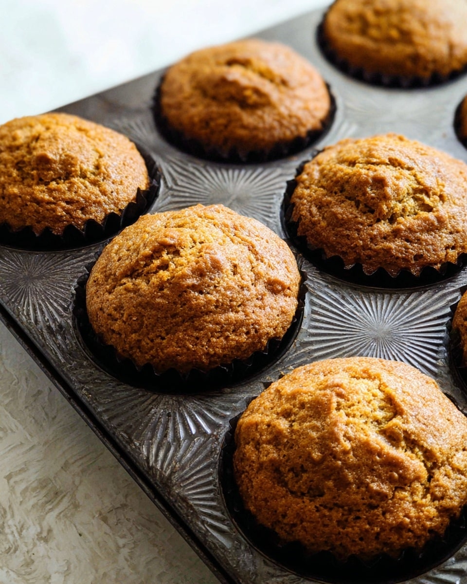 The image shows four brown muffins with a slightly rough top and a textured surface, sitting in dark brown and yellow zigzag-patterned paper cups. They are placed on a white round plate which rests on a cream cloth with subtle wrinkles. Behind the plate is a smooth, small orange pumpkin, all set against a white marbled textured background. photo taken with an iphone --ar 4:5 --v 7