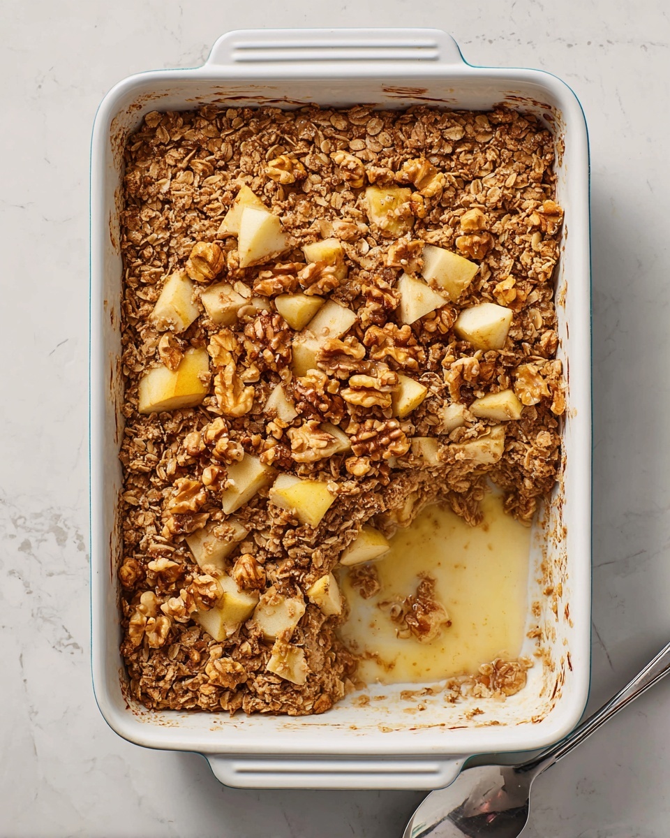 A rectangular white baking dish with a baked oatmeal inside, showing two clear layers: the bottom layer is moist and light brown, and the top layer is a rough, crunchy oat and walnut mix with scattered chunks of pale yellow apple. Part of the oatmeal is scooped out, revealing the smooth texture underneath and some creamy liquid pooling in the empty corner. The oats and walnuts on top have a golden toasted color, and the apple pieces are unevenly placed across the top. The dish rests on a white marbled surface photo taken with an iphone --ar 4:5 --v 7