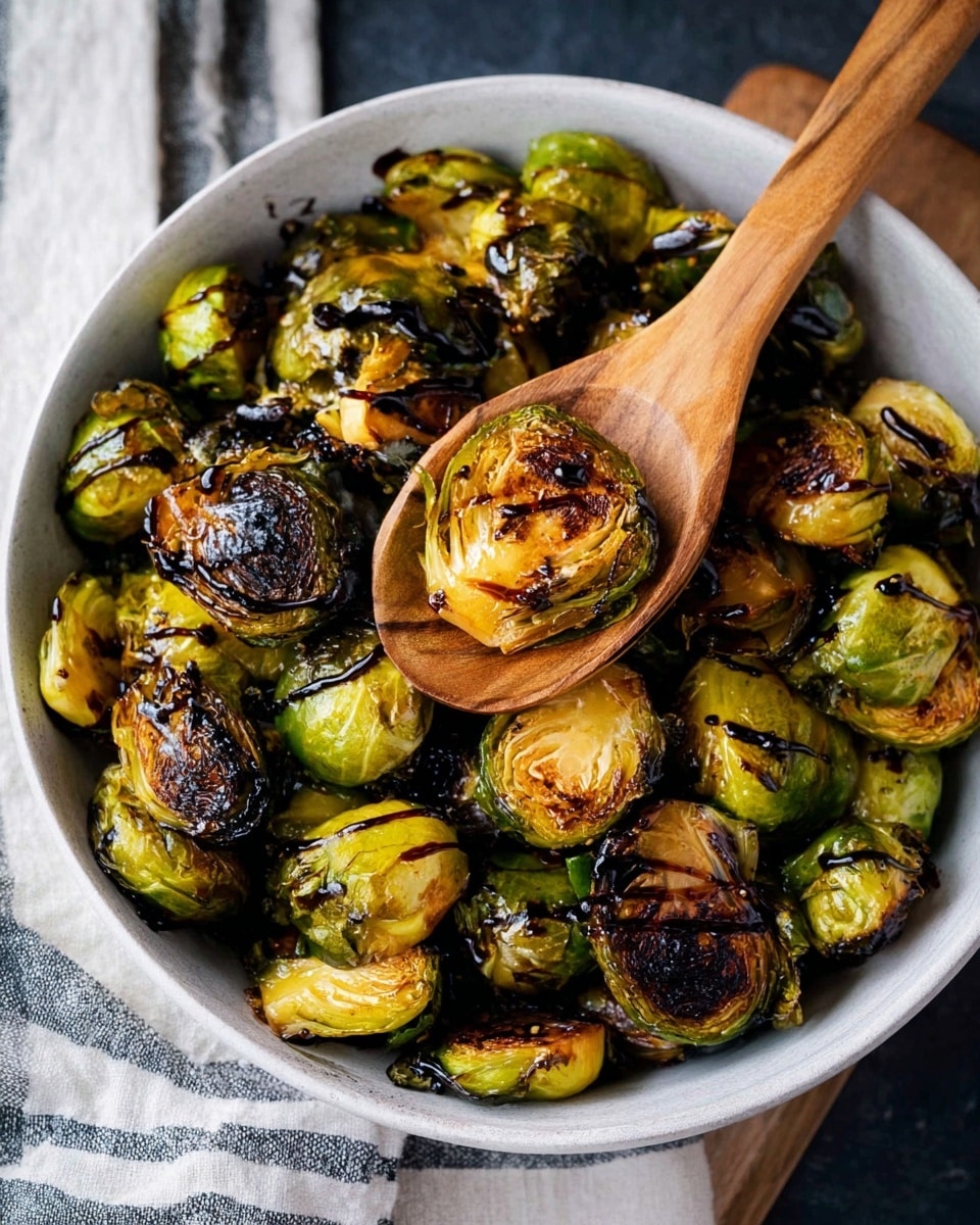 A white square dish filled with roasted Brussels sprouts that are halved, showing a mix of bright green outer leaves and browned, slightly charred centers. The Brussels sprouts have a crispy texture and are piled unevenly, with some layers overlapping. A large silver spoon lies on the right side of the dish, partially submerged in the sprouts. The background is a white marbled surface with a light gray cloth that has soft blue stripes folded casually near the top of the image. Photo taken with an iphone --ar 4:5 --v 7