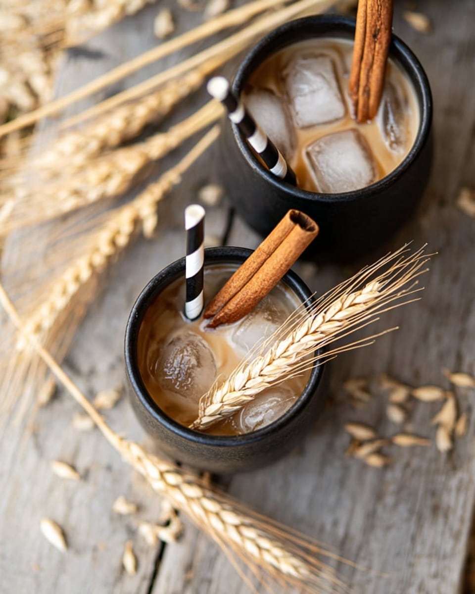 The image shows a top view of two small black cups filled with a light brown creamy drink with ice cubes inside. Each cup has a thick cinnamon stick standing up in the middle and a black and white striped straw on one side. A small bunch of wheat stalks rests next to the straw in the cup in the foreground. The cups are placed on a wooden surface with some scattered wheat stalks around. The background below the wooden surface is a blurry light straw color. photo taken with an iphone --ar 4:5 --v 7