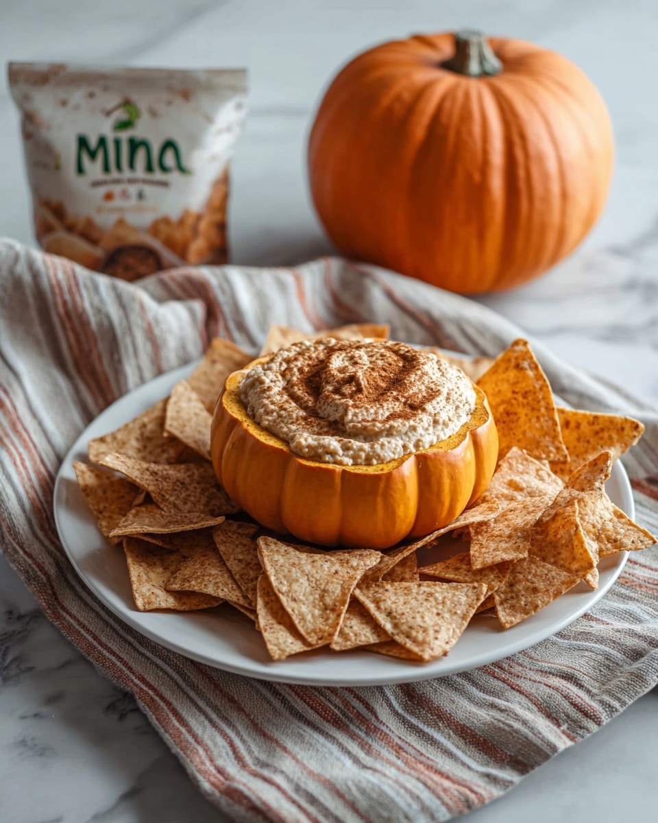 The image shows a bright orange small pumpkin hollowed out and filled with a thick, creamy beige dip sprinkled with dark brown powder on top, creating a textured swirl. The pumpkin sits in the middle of a round white plate filled with many irregularly shaped light brown chips that have a slightly rough texture. The background and surface have a white marbled texture, giving a clean and fresh look. photo taken with an iphone --ar 4:5 --v 7