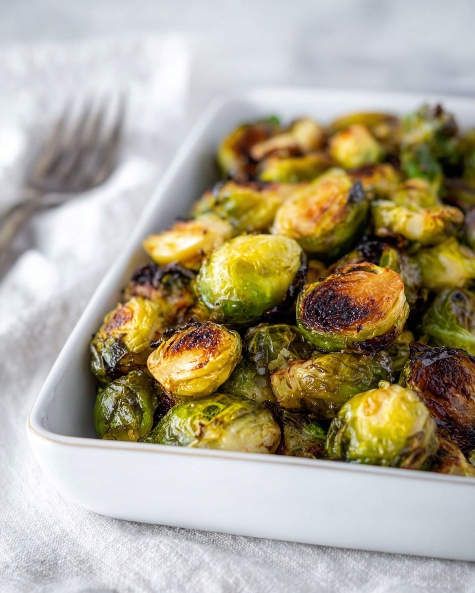 A white rectangular dish filled with roasted Brussels sprouts, each split in half showing a mix of green outer leaves and golden brown, slightly charred centers. The Brussels sprouts have a crispy texture on the browned sides with some still showing vibrant green. The background features a soft-focus white marbled surface with a blurred fork and cloth in the back. The image is close up, focusing on the roasted sprouts inside the dish, creating a warm and appetizing look. photo taken with an iphone --ar 4:5 --v 7