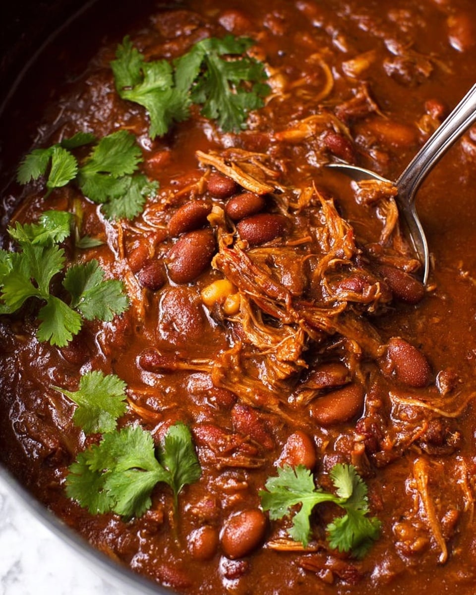 A white bowl full of dark reddish-brown chili with visible pieces of tender shredded meat and red beans. On top, there is a layer of melted orange cheese, followed by a dollop of smooth white sour cream. Fresh green avocado cubes and chopped cilantro leaves sit on the very top, adding bright green color contrast. A silver spoon rests inside the bowl. The bowl is placed on a white marbled surface. Photo taken with an iphone --ar 4:5 --v 7