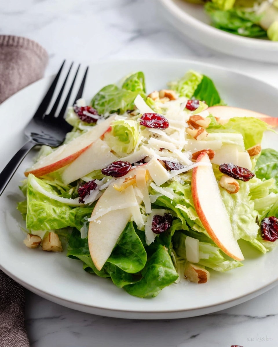 The image shows a wooden bowl filled with fresh green lettuce leaves as the base layer, topped with thin slices of green pears and small cubes of red-skinned apple scattered evenly. There is a light drizzle of creamy white dressing with tiny black seeds on top. The salad is also sprinkled with shredded white cheese, whole cashews, and dried red cranberries adding more color and texture, sitting on a white marbled surface with a blurred apple and a cloth nearby. Photo taken with an iphone --ar 4:5 --v 7
