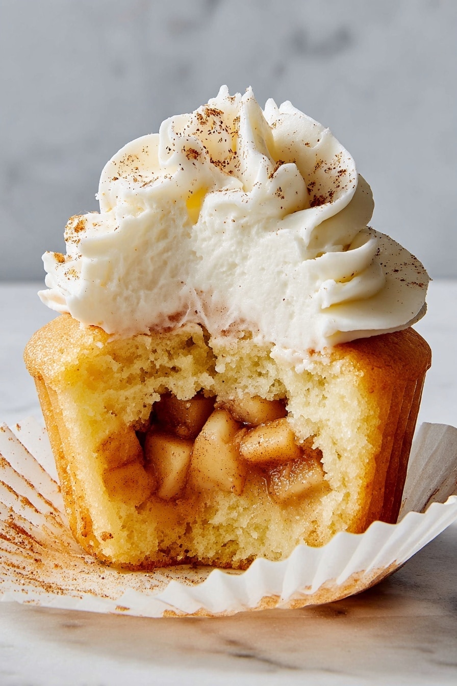 The image shows a cupcake with three visible layers. The bottom layer is a light yellow cake with a soft and moist texture, partially wrapped in a white cupcake liner slightly pulled down. Inside the middle layer, there are small diced apple pieces coated with a cinnamon-colored spice, giving a warm brown shade with a slightly shiny texture. The top layer is a thick swirl of white cream frosting, decorated with small sprinkles of cinnamon spice that add a light brown touch on the smooth creamy surface. The cupcake sits on a white marbled surface. Photo taken with an iphone --ar 2:3 --v 7 - Apple Pie-Stuffed Cupcakes, apple pie cupcakes, apple-filled cupcakes, cinnamon apple cupcakes, fall dessert recipes