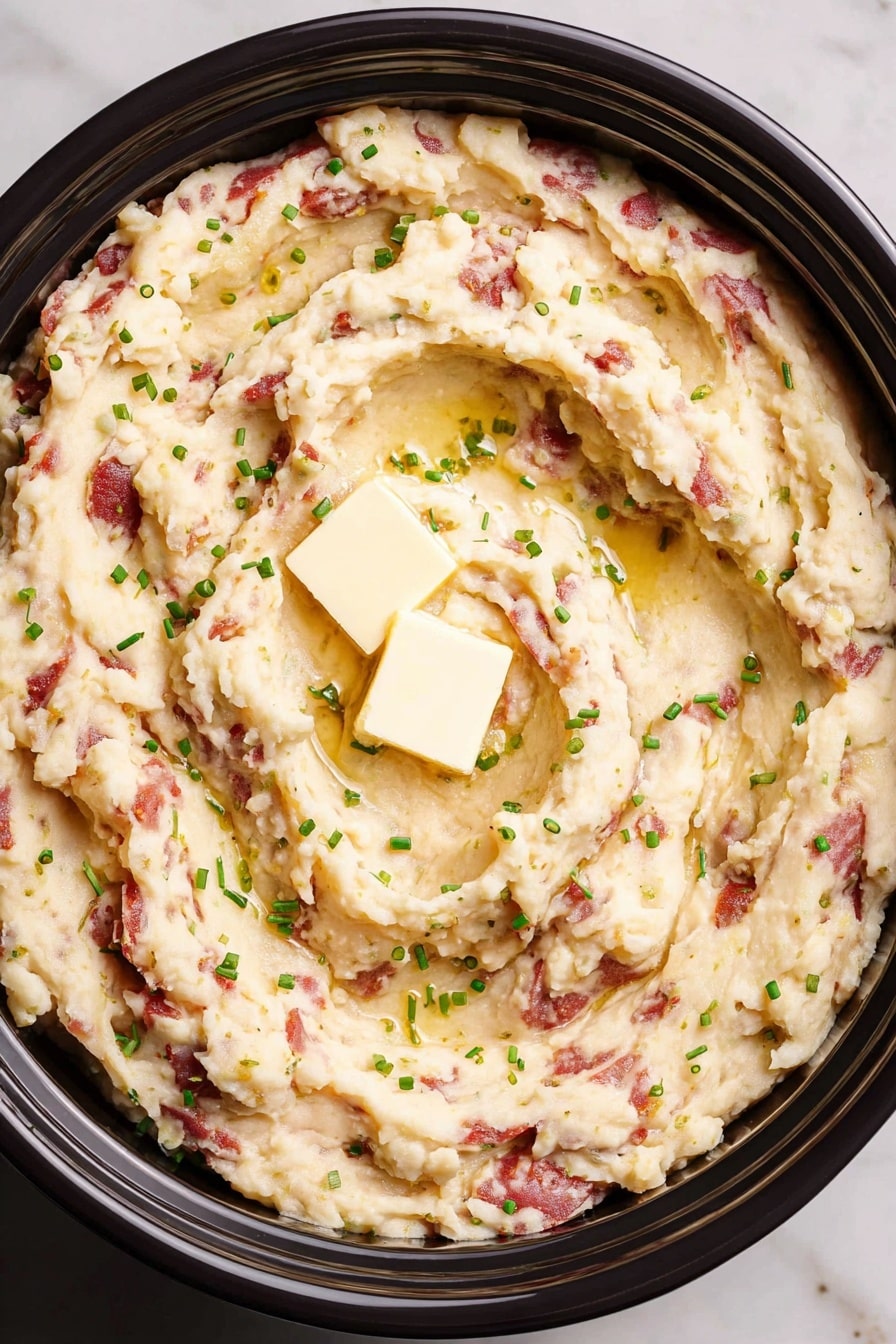 The image shows a close-up of creamy mashed potatoes with chunks of red potato skin spread throughout. The mashed potatoes have a smooth but slightly textured look, with some small lumps and soft peaks. There are small pools of melted butter melting into the potatoes, adding a glossy, light yellow shine. Fresh green chopped chives are sprinkled evenly on top, adding a slight color contrast. The dish is presented in a white bowl against a white marbled background. photo taken with an iphone --ar 2:3 --v 7 - Crockpot Garlic Mashed Potatoes, easy garlic mashed potatoes, slow cooker mashed potatoes, creamy garlic potato recipe, holiday mashed potato side