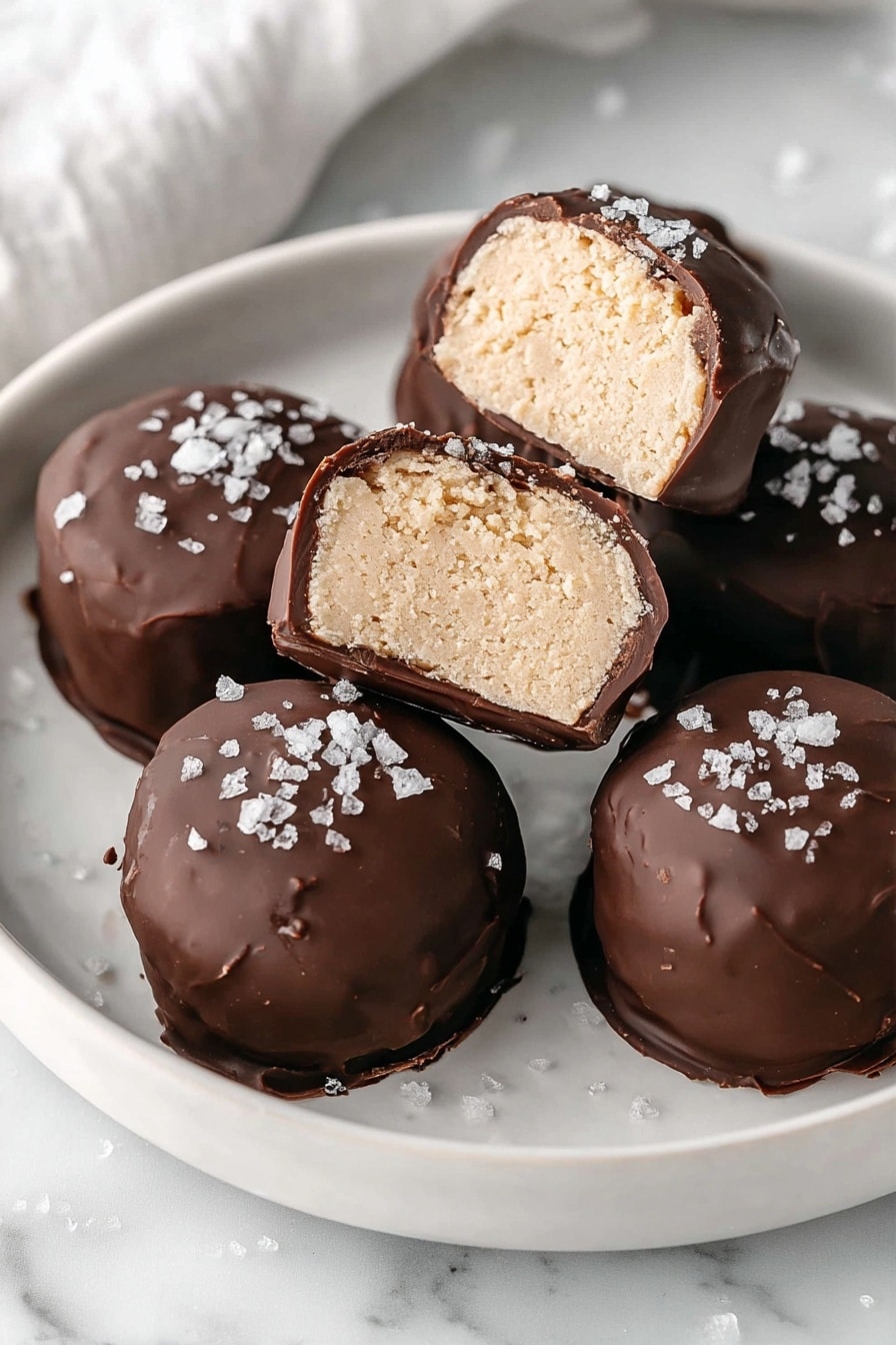 The image shows three round chocolates on a white plate with a white marbled surface. Two chocolates are whole with a smooth, dark chocolate shell sprinkled with coarse white sea salt on top. The third chocolate is cut in half, revealing a thick, creamy beige filling inside with a slightly crumbly texture. The dark chocolate shell forms a smooth, rich outer layer that contrasts with the soft inner filling. Photo taken with an iphone --ar 2:3 --v 7 - High Protein Frozen Peanut Butter Greek Yogurt Bites, healthy protein snacks, easy frozen yogurt bites, nutritious peanut butter dessert, no-bake high protein treat