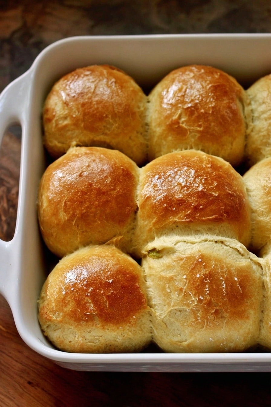 The image shows a close-up of nine round bread rolls inside a white rectangular baking dish with handles, placed closely together. The rolls have a golden brown, shiny, and slightly cracked top crust with a soft texture visible around the edges. The baking dish sits on a wooden surface that is replaced here with a white marbled texture. The lighting is warm, highlighting the golden tones of the bread. photo taken with an iphone --ar 2:3 --v 7 - Sweet Potato and Honey Dinner Rolls, soft sweet potato dinner rolls, homemade sweet potato rolls, honey glazed dinner rolls, easy sweet potato bread