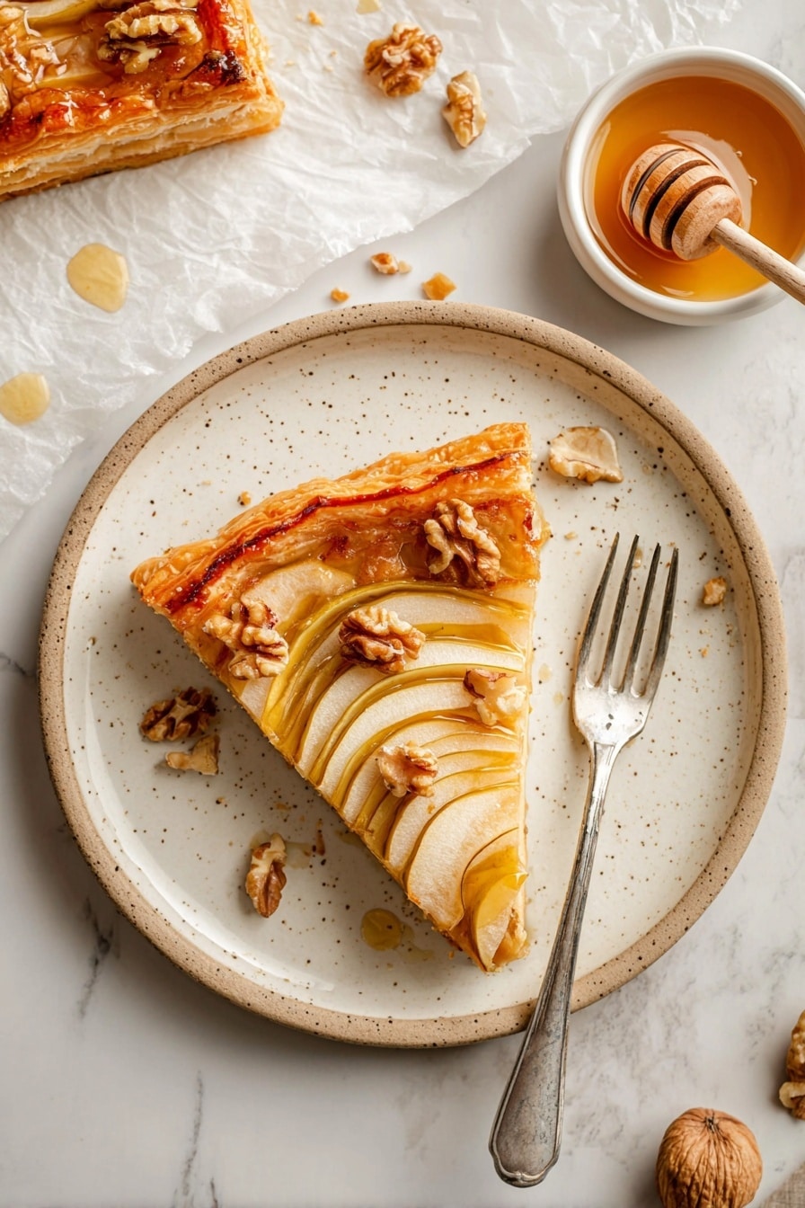 The image shows a white speckled plate with a slice of golden puff pastry tart on it. The tart has a thick, crispy outer crust with a shiny finish. Inside, there is a layer of thinly sliced pear arranged in a fan shape and topped with small chunks of walnuts. The pear slices have a soft beige color with a slight translucence. Some honey glaze is drizzled over the tart, adding a glossy shine. A silver fork rests on the right side of the plate. In the background, there is a small white bowl with light amber honey and a wooden honey dipper inside it, along with a few scattered walnut pieces and a corner of another tart slice on white parchment paper. The whole setting is on a white marbled surface. photo taken with an iphone --ar 2:3 --v 7 - Pear, Walnut & Cheddar Puff Pastry Tart, savory pear and cheese tart, quick puff pastry tart recipe, easy cheese and fruit tart, cozy dinner party tart