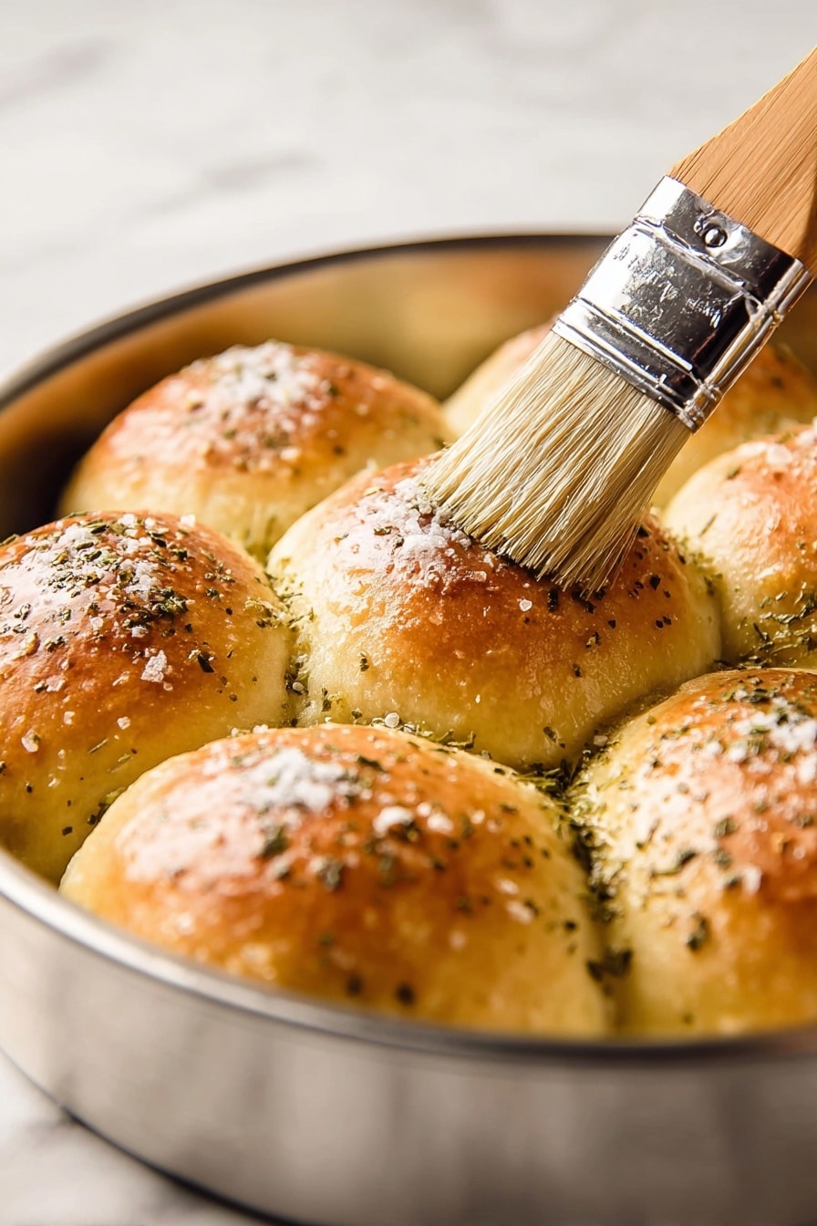 A round baking pan holds eight golden brown dinner rolls arranged close together in a single layer. Each roll has a smooth, shiny top sprinkled with small white salt flakes and greenish dried herbs. A wooden brush with soft bristles is brushing a shiny glaze over the top of one roll, adding a moist, glossy texture. The pan edges are visible, showing a metallic surface. The background is a white marbled texture. photo taken with an iphone --ar 2:3 --v 7 - Herby Rhodes Rolls, easy herb bread, fluffy herb rolls, buttery herb dinner rolls, homemade herbed bread