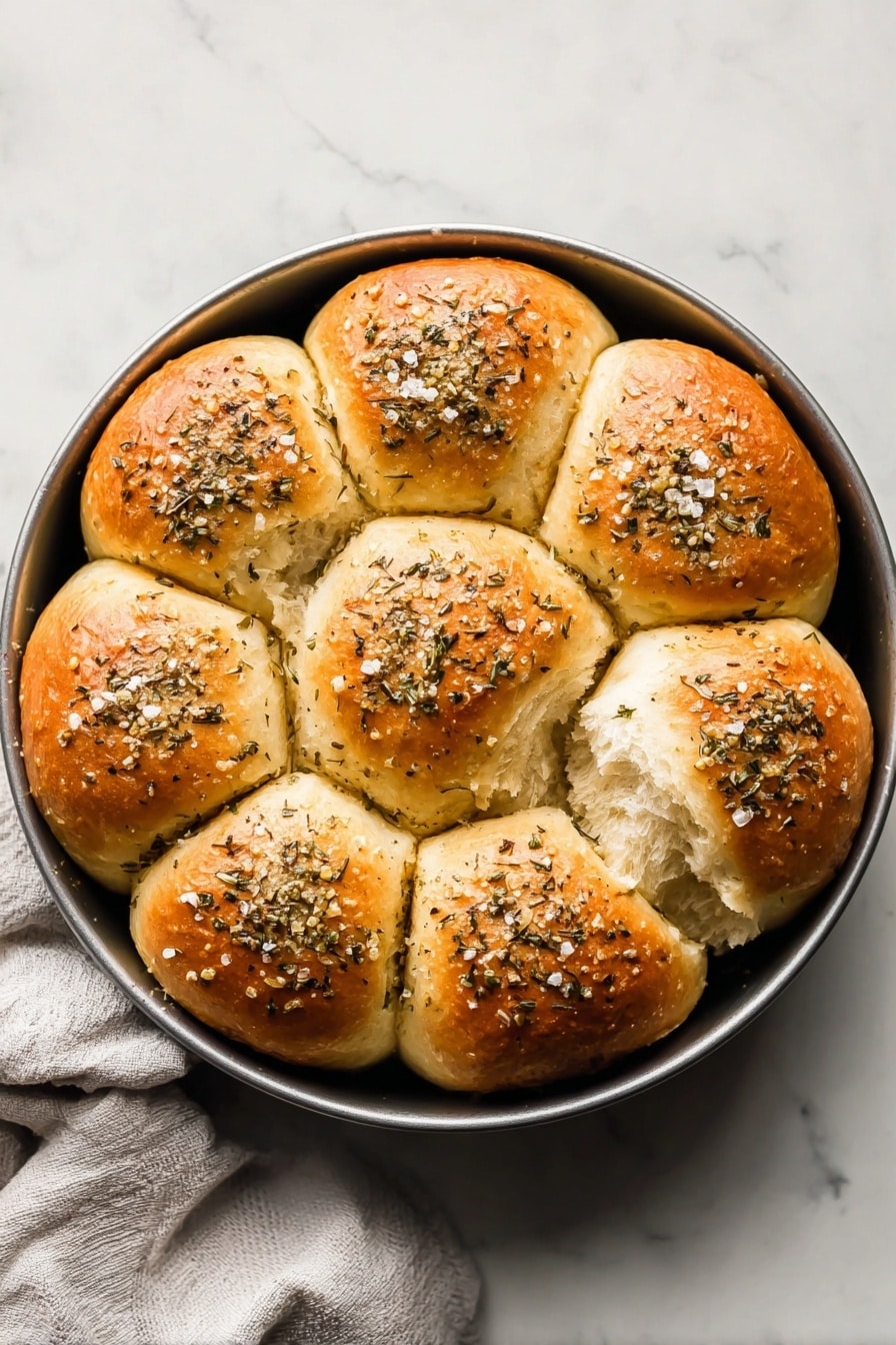 This image shows a round pan filled with nine golden-brown dinner rolls arranged in a single layer. Each roll is soft with a shiny top sprinkled generously with dried herbs and coarse salt, giving a textured appearance. The rolls are touching each other, except one roll on the right side that looks slightly pulled away, exposing the fluffy and light inside crumb. The pan is placed on a white marbled surface with a light gray cloth partially visible at the bottom left. Photo taken with an iphone --ar 2:3 --v 7 - Herby Rhodes Rolls, easy herb bread, fluffy herb rolls, buttery herb dinner rolls, homemade herbed bread