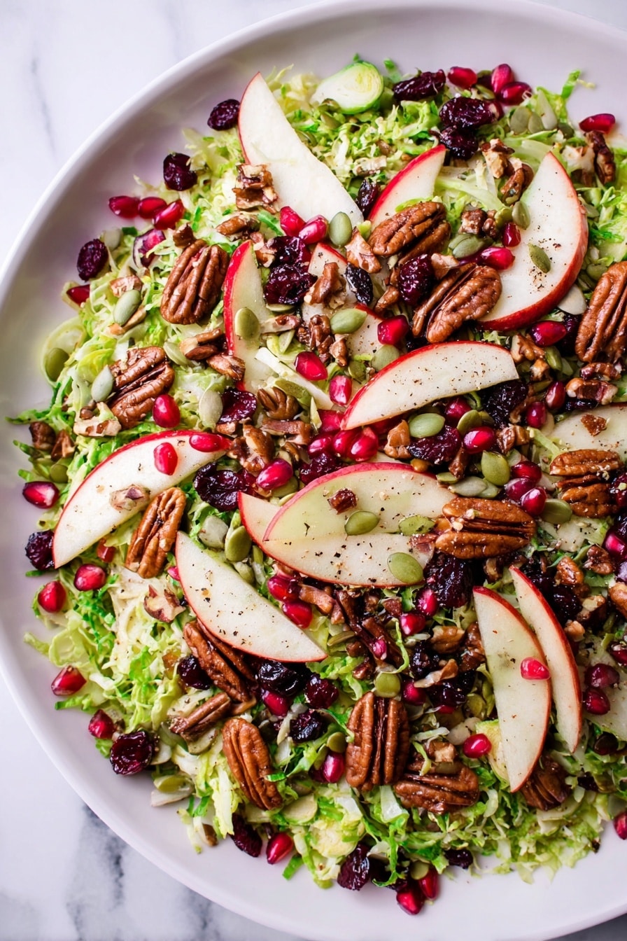 A white bowl filled with a fresh salad showing several layers, the bottom layer is shredded green Brussels sprouts, giving a leafy and slightly rough texture. On top, there are thin, round slices of red-skinned apple spread evenly around the bowl, adding a smooth and shiny look. Scattered over the salad are whole brown pecans, dark red dried cranberries, and small green pumpkin seeds, providing varied textures and rich colors. Bright red pomegranate seeds are sprinkled throughout, adding small pops of color. The salad appears lightly seasoned with tiny specks of black pepper visible. The white bowl rests on a white marbled surface, completing a clean and fresh presentation. photo taken with an iphone --ar 2:3 --v 7 - Shaved Brussels Sprout Salad with Creamy Maple Dressing, Brussels sprout salad, healthy Brussels sprout recipes, winter salad ideas, easy vegetable salads