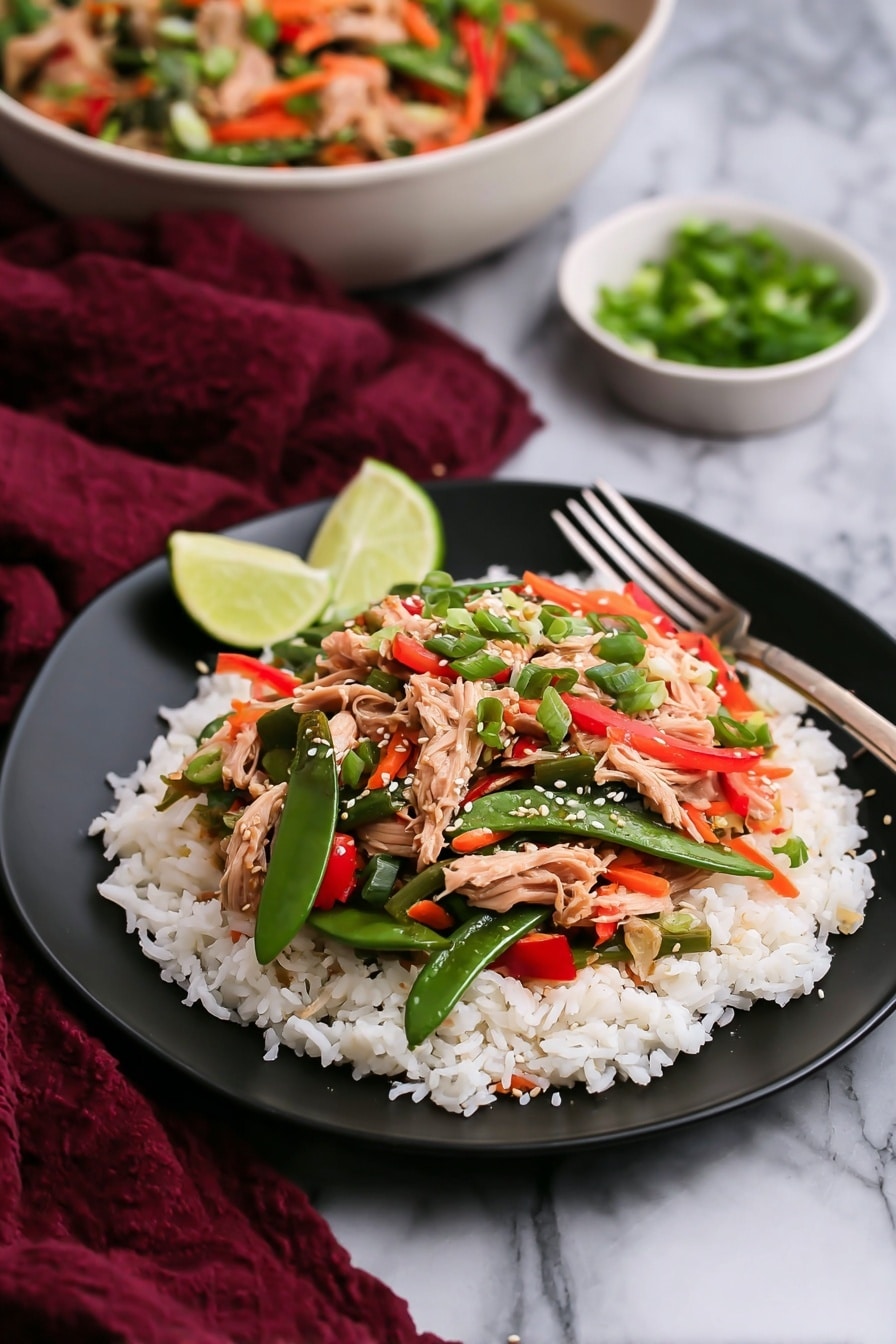 A black plate holds a dish with two clear layers: the bottom layer is soft white rice spread evenly, and the top layer is a colorful mix of cooked vegetables and light pink shredded meat, including bright green snap peas, thin orange carrot strips, red pepper slices, and green scallions, sprinkled with light sesame seeds. On the side of the plate is a small light green lime wedge. In the background, there is a white bowl filled with the same vegetable and meat mix, with a spoon resting inside, and a small white bowl with chopped green onions. The whole setup is on a white marbled surface with a dark red cloth draped casually nearby. Photo taken with an iphone --ar 2:3 --v 7 - Leftover Turkey Stir Fry with Thai Basil and Veggies, turkey leftover stir fry, easy turkey stir fry recipe, Thai basil stir fry, quick leftover turkey meal