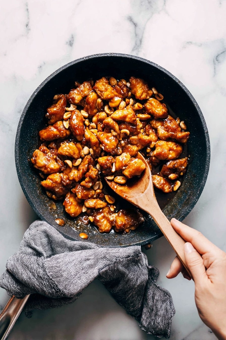 A dark pan filled with many pieces of golden brown cooked chicken mixed with light brown peanuts all coated in a shiny rich sauce, a wooden spoon is lifting some pieces from the pan while a woman's hand is holding the pan's handle wrapped with a grey cloth, all on a white marbled surface photo taken with an iphone --ar 2:3 --v 7 - Cashew Chicken Stir-Fry, easy chicken stir-fry, quick dinner recipes, healthy Asian stir-fry, flavorful chicken and cashews