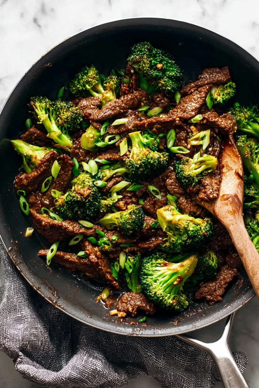 A black skillet filled with stir-fried beef slices and bright green broccoli florets, mixed with small bits of toasted garlic and topped with thin, fresh green onion slices scattered across the dish. A wooden spatula rests inside the skillet, partially lifting some of the food. The skillet sits on a gray textured cloth on a white marbled surface. The beef is dark brown and glossy from the sauce, while the broccoli is vibrant and tender. Photo taken with an iphone --ar 2:3 --v 7 - Sesame Beef and Broccoli Stir Fry, quick beef stir fry, healthy Asian stir fry, easy beef and broccoli recipe, flavorful stir fry dinner