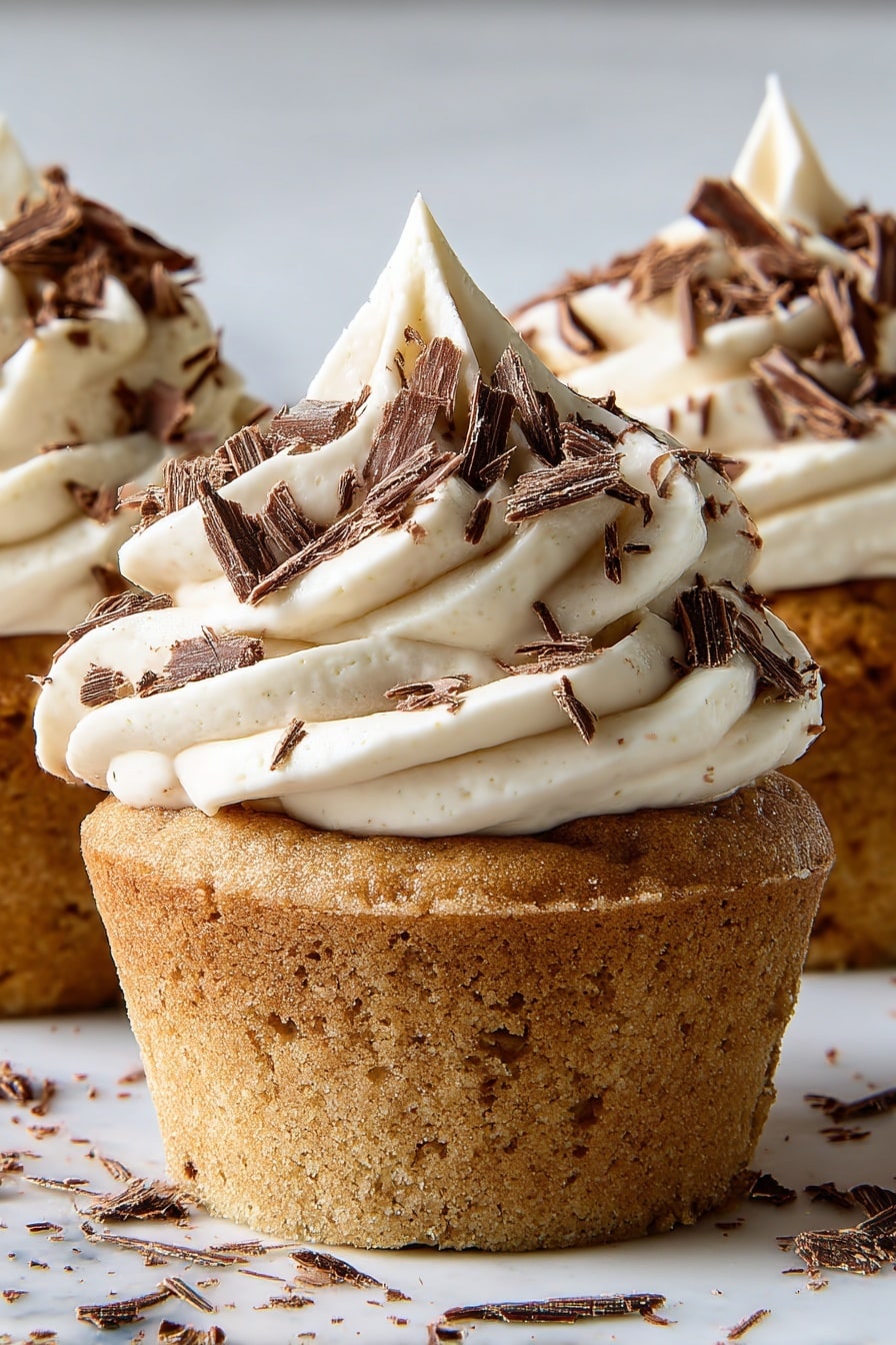 The image shows several cupcakes arranged closely together on a white marbled surface. Each cupcake has two layers: a dense, golden-brown cake base and a thick swirl of light beige cream frosting on top. The frosting is piped in a spiral pattern, rising to a peak in the center. Dark chocolate shavings are scattered generously over the frosting, adding texture and contrast. The cupcakes are well-lit, showing the soft texture of the frosting and the crumbly surface of the cake. Photo taken with an iphone --ar 2:3 --v 7 - White Russian Cupcakes, White Russian Cocktails, Kahlúa Cupcakes, Boozy Cupcake Recipes, Easy Cocktail-Inspired Dessert