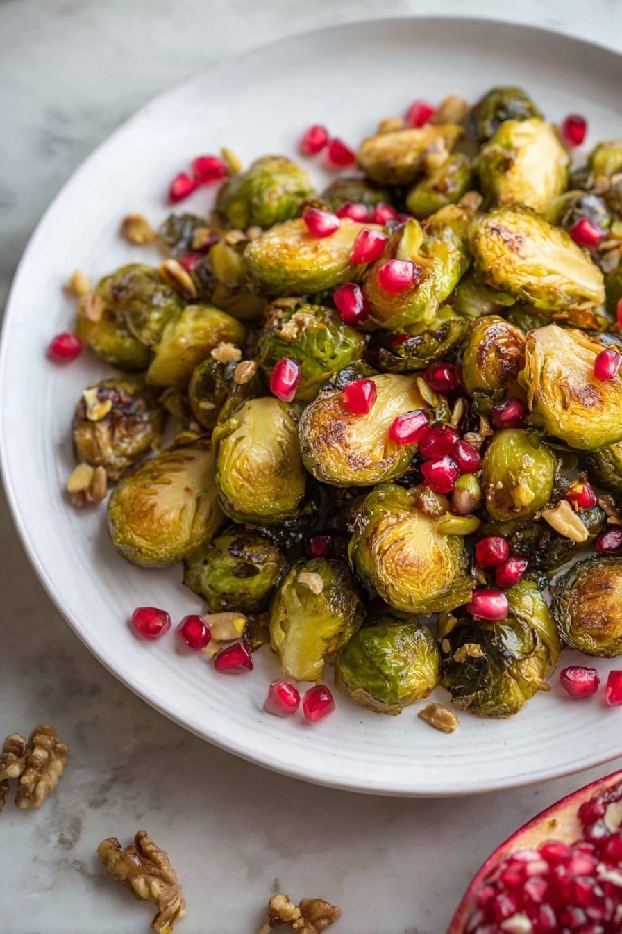 A white plate holds a pile of roasted Brussels sprouts, some whole and some cut in halves, showing a golden brown and slightly crispy texture. Scattered evenly on top and around the sprouts are bright red pomegranate seeds and small greenish-brown nuts, adding color and texture contrast. The plate sits on a white marbled background, and in the lower right corner, half of a pomegranate fruit is visible, showing vibrant red seeds inside. Photo taken with an iphone --ar 2:3 --v 7 - Roasted Maple Mustard Brussels Sprouts with Pistachios and Pomegranate, Brussels sprouts with maple mustard, festive veggie side dish, healthy holiday Brussels sprouts, easy roasted Brussels sprouts