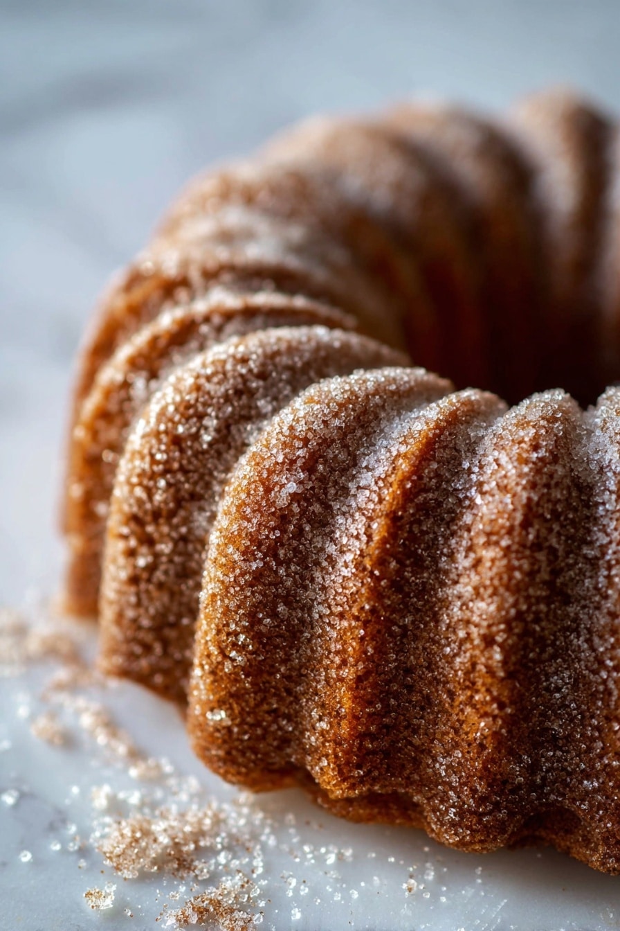 A single brown bundt cake sits in the middle of a white marbled surface, dusted evenly with fine white sugar that highlights the curved ridges of the cake. The cake’s shape is round with deep, symmetrical grooves running vertically around the edges and an empty circular center. The texture of the cake appears soft yet slightly firm, with a light coating of sugar granules creating a sparkling effect against the warm brown color. Photo taken with an iphone --ar 2:3 --v 7 - Apple Cider Donut Cake with Brown Butter and Cinnamon Sugar, Fall dessert recipes, Cozy apple spice cake, Homemade apple cider cake, Easy seasonal cake recipes