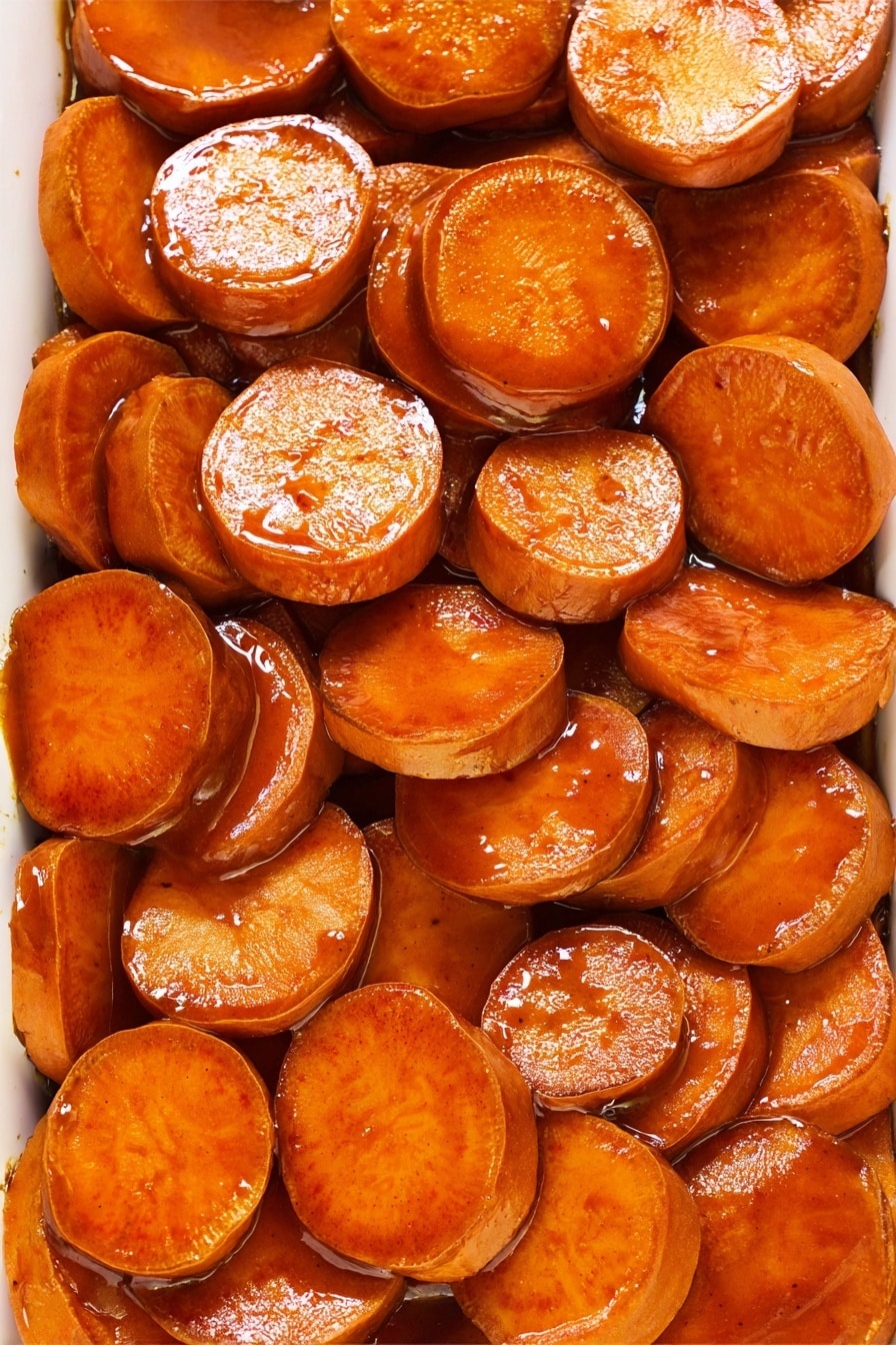 A rectangular white baking dish filled with several layers of thick, round sweet potato slices covered in a shiny brown sauce. The sweet potato slices are a warm orange color with a slightly glossy texture, stacked unevenly but covering the entire dish. To the side, three whole sweet potatoes rest on a striped cloth with brown and white lines, all placed on a white marbled surface. The bright natural lighting highlights the glossy sauce and the vibrant color of the slices, giving the dish a fresh and inviting look. photo taken with an iphone --ar 2:3 --v 7 - Candied Sweet Potatoes, Sweet Potatoes Side Dish, Caramelized Sweet Potatoes, Holiday Sweet Potato Recipe, Cozy Sweet Potato Ideas