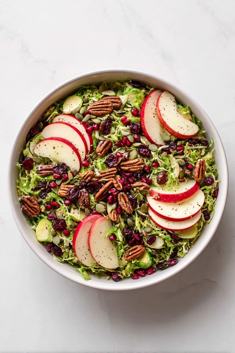 A white bowl is filled with a bright salad consisting of finely shredded green Brussels sprouts as the base layer, topped with thin slices of red and white apple arranged in groups around the bowl. Dark brown pecans and scattered red pomegranate seeds add texture and color contrast, along with small green pumpkin seeds spread evenly. There are also dark dried cranberries distributed throughout. The salad is lightly sprinkled with black pepper. Next to the bowl, there is a small white sauce dish filled with a creamy light brown dressing set on a white marbled surface. photo taken with an iphone --ar 2:3 --v 7 - Shaved Brussels Sprout Salad with Creamy Maple Dressing, Brussels sprout salad, healthy Brussels sprout recipes, winter salad ideas, easy vegetable salads