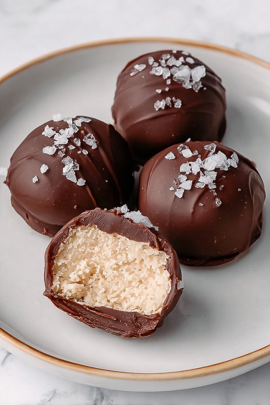 The image shows five round chocolate-covered treats on a white plate, each with a smooth dark brown outer layer. Two of the treats are cut in half to reveal a creamy, light beige filling with a dense texture inside. The chocolate coating is thin and shiny, with a few flakes of white sea salt sprinkled on top, adding contrast. The plate sits on a white marbled surface. photo taken with an iphone --ar 2:3 --v 7 - High Protein Frozen Peanut Butter Greek Yogurt Bites, healthy protein snacks, easy frozen yogurt bites, nutritious peanut butter dessert, no-bake high protein treat