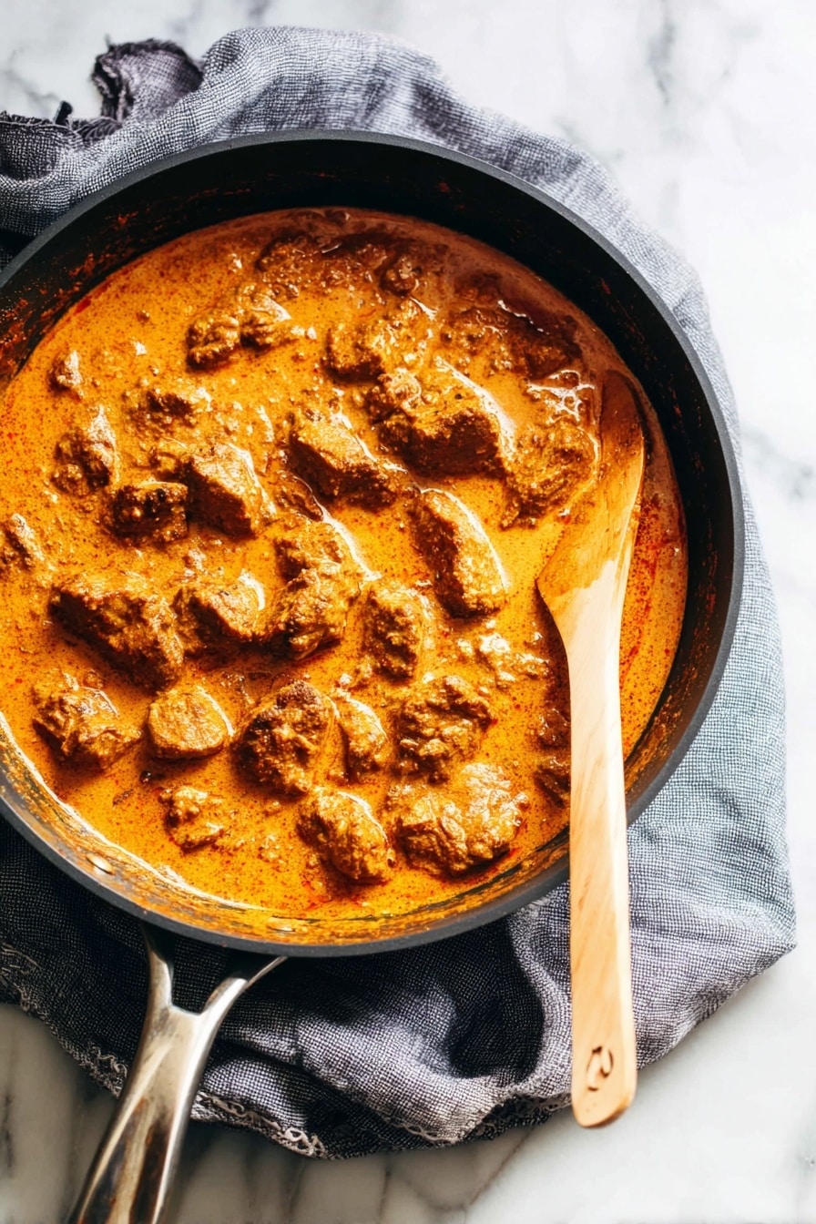 A dark round pan filled with a thick orange sauce that covers many uneven pieces of browned meat. The sauce looks smooth and creamy, with some darker spots where it has cooked more. A light wooden spoon with sauce on it rests on the right edge of the pan, partly submerged. The pan sits on a gray cloth on a white marbled surface. Photo taken with an iphone --ar 2:3 --v 7 - Easy Chicken Tikka Masala, Chicken Tikka Masala, homemade Chicken Tikka Masala, simple Indian curry, quick Chicken Tikka Masala