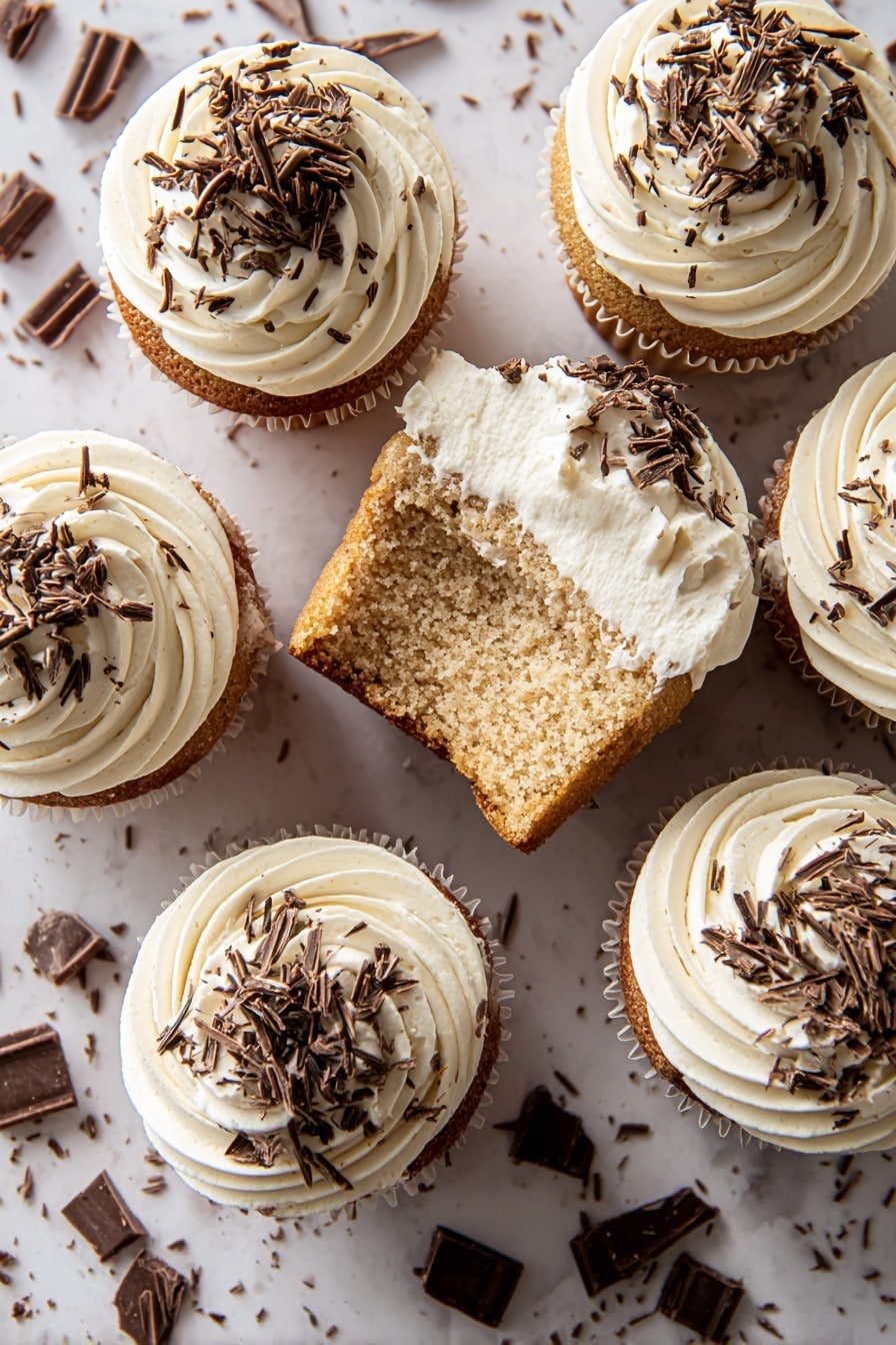 The image shows seven cupcakes on a white marbled surface. Each cupcake has two layers: the bottom layer is a soft, light brown cake with a slightly rough texture, and the top layer is a thick swirl of creamy white frosting. The frosting is decorated with small, thin pieces of dark chocolate sprinkled all over. One cupcake is shown from the side with a bite taken out, revealing the clear division between the cake and frosting layers. The cupcakes are arranged in a close group with chocolate pieces scattered around them. photo taken with an iphone --ar 2:3 --v 7 - White Russian Cupcakes, White Russian Cocktails, Kahlúa Cupcakes, Boozy Cupcake Recipes, Easy Cocktail-Inspired Dessert