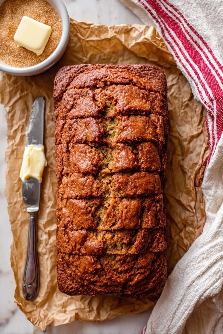 A loaf of sliced banana bread with a dark golden brown crust is placed on crinkled parchment paper. The banana bread has a rough, textured surface with visible cracks and a dense, moist interior. To the left of the bread, there is a white bowl filled with light brown sugar, and a metal knife with a pat of pale yellow butter resting on the blade. A white kitchen towel with red stripes is draped on the right side of the image, resting on a white marbled surface. Photo taken with an iphone --ar 2:3 --v 7 - Moist Banana Bread, moist banana bread, banana bread recipe, soft banana bread, easy banana bread
