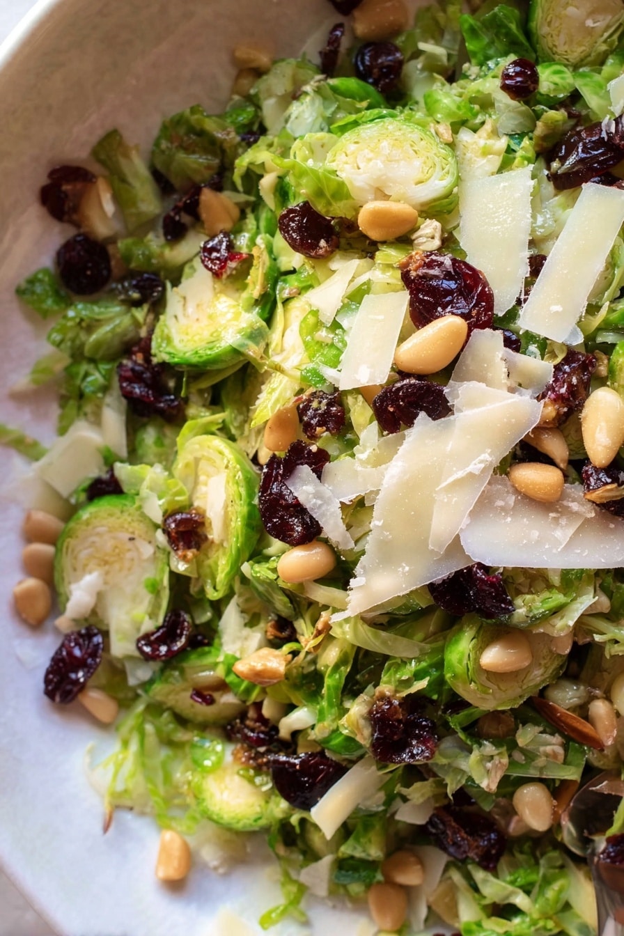 A white bowl filled with a fresh salad made of thinly sliced bright green Brussels sprouts as the base layer, mixed with scattered dark red dried cranberries and light brown toasted pine nuts, topped with thin, uneven flakes of pale yellow cheese, all sprinkled with black pepper. Two silver utensils, a spoon on the left and a fork leaning on the right side of the bowl, rest inside it. Nearby, there is a small glass bowl with a golden dressing and a spoon in it, and some cheese flakes scattered around the white marbled surface. A white cloth with light brown stripes is partly visible in the top left corner. Photo taken with an iphone --ar 2:3 --v 7 - Brussels Salad with Spiced Maple Vinaigrette, Brussels sprout salad, Maple vinaigrette salad, healthy Brussels sprout dish, easy Brussels salad recipe