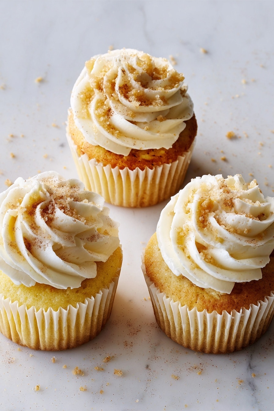 The image shows a group of yellow cupcakes on a white plate, each topped with fluffy cream-colored frosting swirled into soft peaks and sprinkled with light brown cinnamon powder. One cupcake in the front has its white paper wrapper peeled down and a bite taken out of it, revealing a filling of small, caramel-colored apple chunks mixed with cinnamon inside the soft yellow cake. The background is a white marbled surface. The lighting is soft, bringing out the creamy and moist texture of the cupcakes. photo taken with an iphone --ar 2:3 --v 7 - Apple Pie-Stuffed Cupcakes, apple pie cupcakes, apple-filled cupcakes, cinnamon apple cupcakes, fall dessert recipes