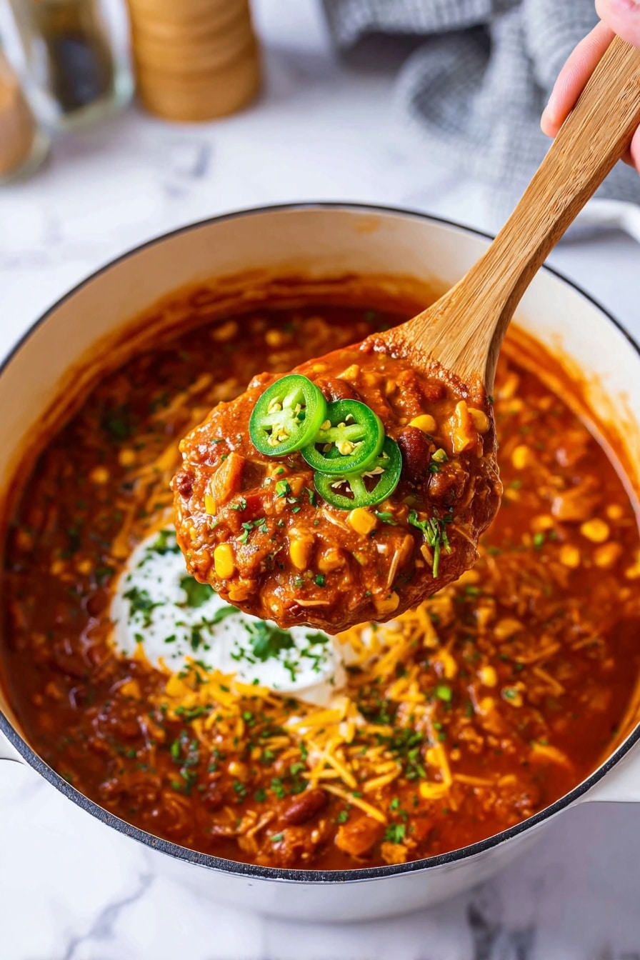 A white bowl filled with thick, chunky chili that has visible pieces of corn and beans in a rich red sauce. On top, there is a generous dollop of white sour cream layered with four bright green jalapeño slices and sprinkles of chopped fresh green herbs. The bowl sits on a blue cloth over a white marbled surface, with blurred salt and pepper shakers and a green pepper in the background, and a glass of iced amber drink to the side. photo taken with an iphone --ar 2:3 --v 7 - Leftover Turkey Chili, turkey leftover recipes, hearty turkey chili, easy leftover turkey dinner, flavorful turkey chili