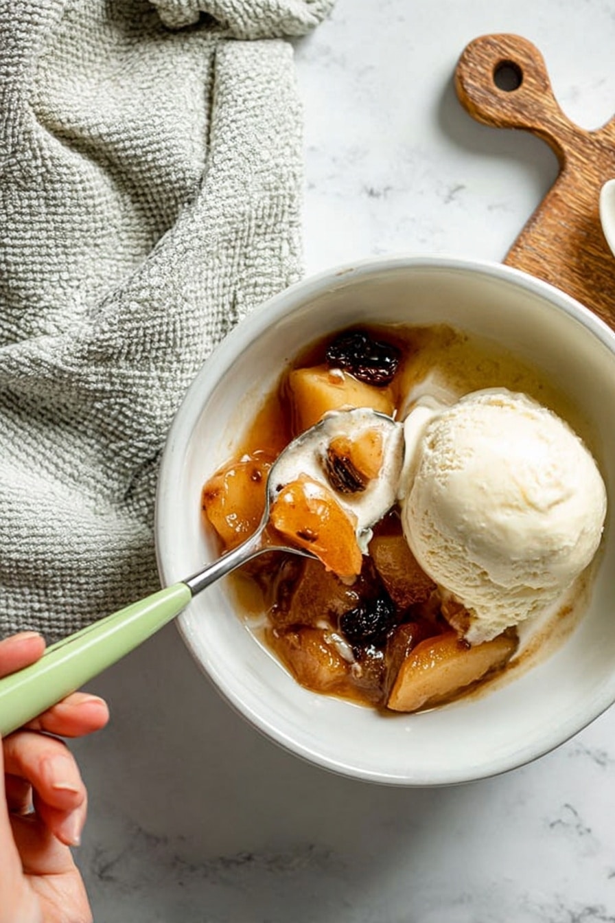 The image shows a white bowl filled with a warm dessert made of soft cooked fruit pieces in a brown syrup, topped with a smooth scoop of vanilla ice cream melting slightly on one side. A spoon with a light green handle is held by a woman's hand, lifting a bite that includes a piece of fruit with syrup and a dark raisin. The bowl sits on a white marbled surface, with a light gray textured cloth and a small wooden cutting board placed near the top edge. The overall tone is warm and cozy, with natural lighting highlighting the creamy and syrupy textures. photo taken with an iphone --ar 2:3 --v 7 - Baked Apple, baked apple dessert, easy baked apple recipe, cozy apple dessert, comforting baked apple