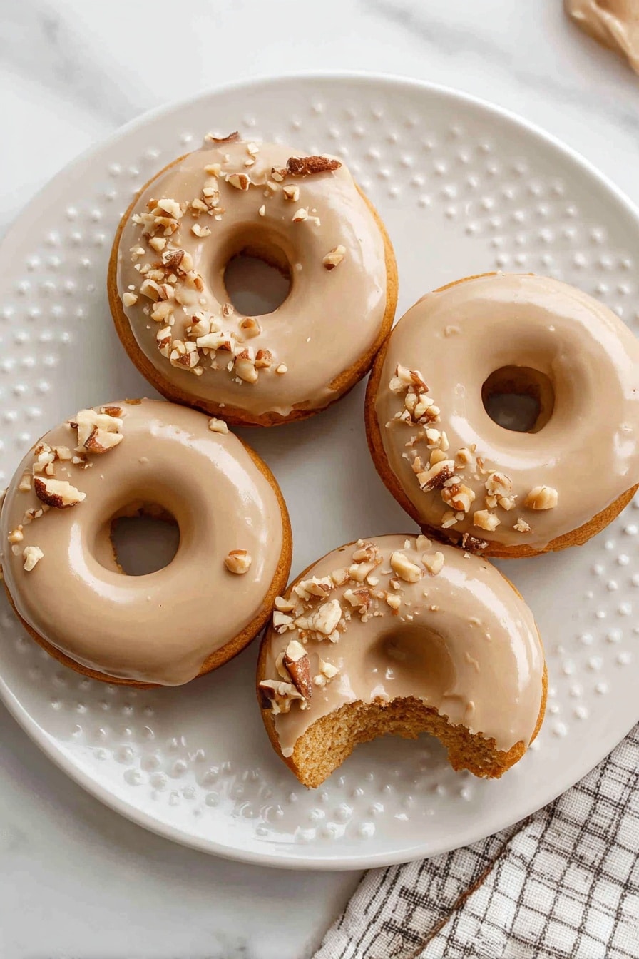 There are four round donuts on a white plate with a light dotted pattern. Each donut has a smooth, light brown glaze covering the top layer, with small pieces of chopped nuts sprinkled over the glaze. One donut shows a bite taken out of it, revealing a soft, orange-brown inside layer. The plate is placed on a white marbled surface and part of a checkered cloth is visible in the corner. Photo taken with an iphone --ar 2:3 --v 7 - Gluten-Free Pumpkin Donuts, Pumpkin Donuts with Cinnamon Sugar, Pumpkin Donuts with Brown Sugar Glaze, Fall Pumpkin Donut Recipe, Gluten-Free Fall Treats
